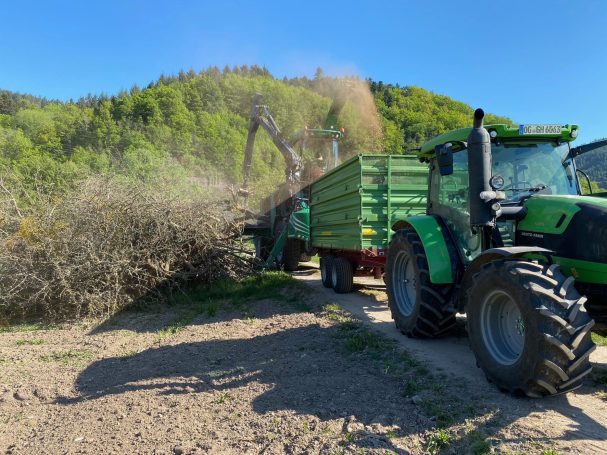 Häckseln für unsere Hackschnitzel-Heizung Ein Traktor mit Anhänger zerkleinert Äste vor bewaldetem Hang bei sonnigem Wetter.
