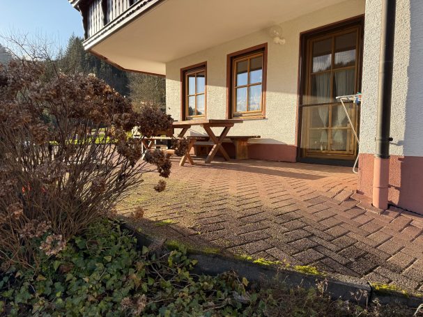 Große Terrasse Terrasse mit Holzbank und Tisch, umgeben von Pflanzen und einem gepflasterten Boden.