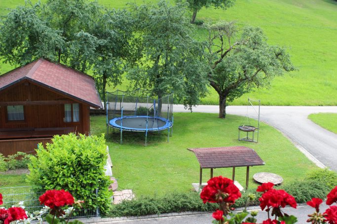 Grillplatz mit Sandkasten und Trampolin Grüner Garten mit Trampolin, Holzhaus und blühenden roten Blumen im Vordergrund.