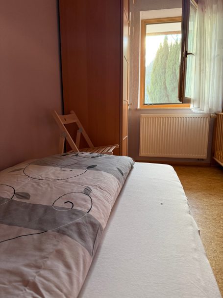 Kinderzimmer Ein einfaches Zimmer mit einem Bett, einem Fenster und einem Stuhl in der Ecke.