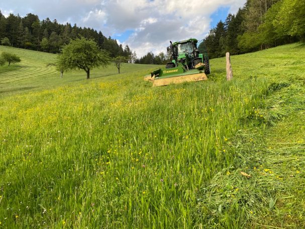 Traktor mit Mähwerk Traktor mäht Gras auf einer grünen Wiese mit Bäumen im Hintergrund.