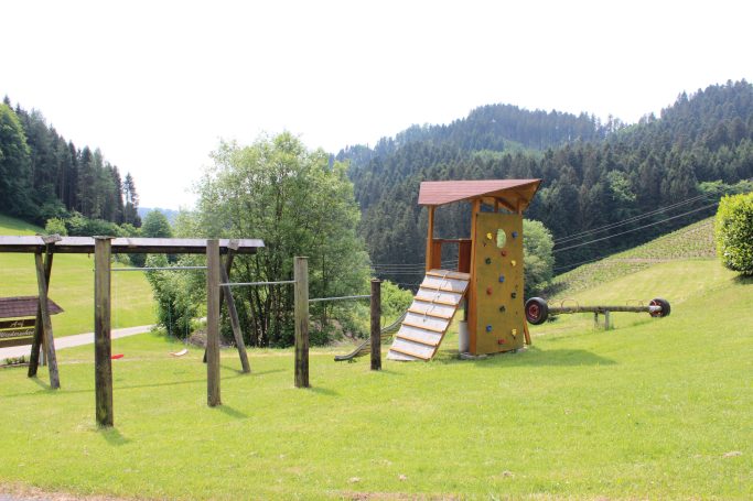 Spielplatz Spielplatz mit Kletterturm, Schaukeln und Rasenfläche, umgeben von Bäumen und Hügeln.