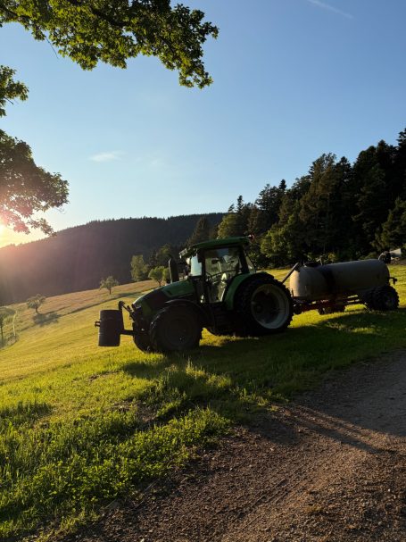 Die Felder düngen Grüner Traktor auf einer Wiese vor einer bewaldeten Hügelkulisse bei Sonnenuntergang.